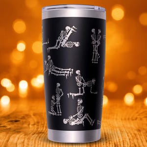 May include: Black stainless steel tumbler with a silver rim and lid. The tumbler features a repeating pattern of white skeleton figures in various poses. The background is a blurred orange and brown.