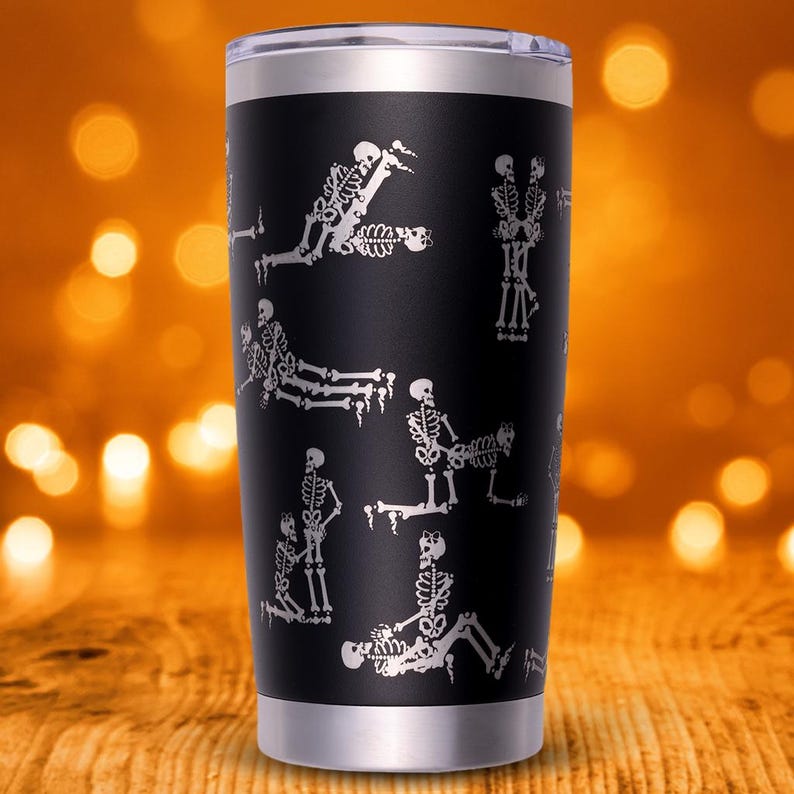 May include: Black stainless steel tumbler with a silver rim and lid. The tumbler features a repeating pattern of white skeleton figures in various poses. The background is a blurred orange and brown.