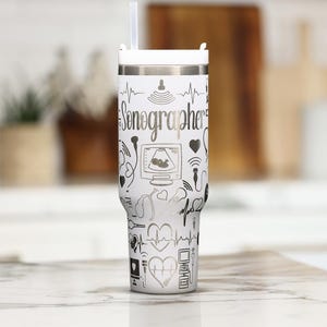 Puede incluir: Un vaso de acero inoxidable blanco con tapa blanca y pajita transparente. El exterior presenta un patrón repetitivo de iconos de líneas negras relacionados con la ecografía, incluyendo monitores, corazones y sondas, con la palabra Sonographer.