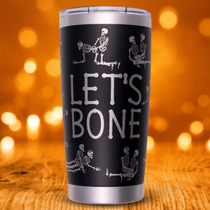 May include: Black stainless steel tumbler with the phrase "LET'S BONE" in white lettering. The tumbler features skeleton illustrations around the cup. The tumbler has a silver rim and a clear lid. The background is blurred with orange and yellow bokeh lights.