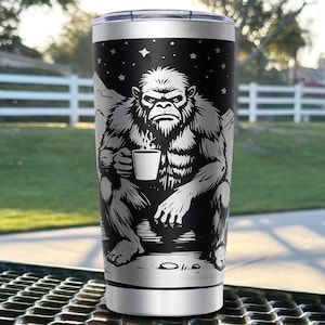 May include: A stainless steel tumbler featuring a black and white graphic of a seated Bigfoot creature holding a steaming mug of coffee. The design includes a starry night sky background with mountain silhouettes, topped with a clear plastic lid.
