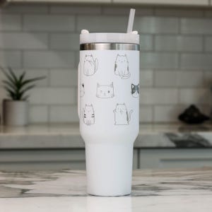 Puede incluir: Un vaso de acero inoxidable blanco con tapa negra y una pajita. El vaso tiene un dise&ntilde;o de varios gatos en diferentes poses.