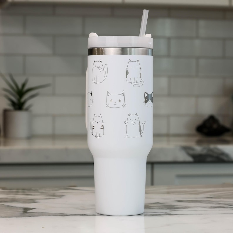 Puede incluir: Un vaso de acero inoxidable blanco con tapa negra y una pajita. El vaso tiene un dise&ntilde;o de varios gatos en diferentes poses.