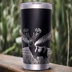 Könnte beinhalten: Ein mattschwarzer Edelstahlbecher mit einer lasergravierten Grafik eines Hirsches, der auf einer Wiese vor einem Wald und einer Bergkette steht. Das zylindrische Gefäß verfügt über einen silberfarbenen Rand sowie einen passenden silbernen Boden.