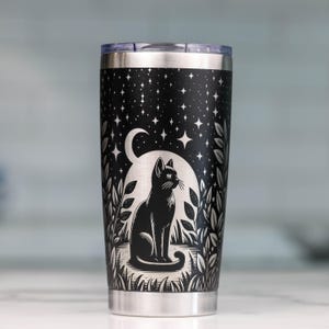 May include: A black stainless steel tumbler featuring a laser-engraved design of a cat sitting in a field of plants under a starry night sky with a large crescent moon. The cylindrical vessel has a metallic silver rim and a matching base.