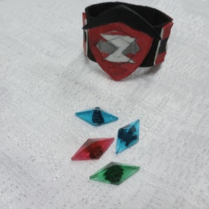 May include: A black and red wristband with a shield design and four colorful, diamond-shaped gems. The gems are blue, red, and green. The wristband has a red shield with a white and gray design.