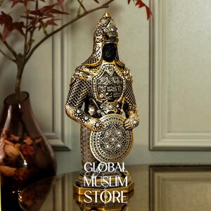 May include: A gold and black decorative figurine of a warrior holding a shield. The warrior is wearing a helmet and armor with intricate details. The shield is decorated with a geometric pattern and jewels.