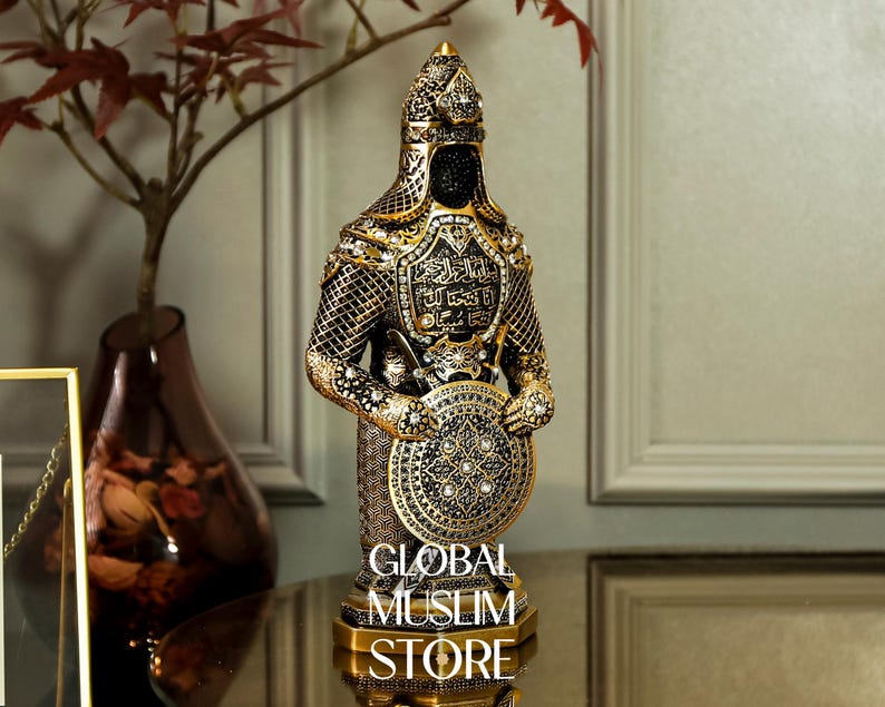 May include: A gold and black decorative figurine of a warrior holding a shield. The warrior is wearing a helmet and armor with intricate details. The shield is decorated with a geometric pattern and jewels.