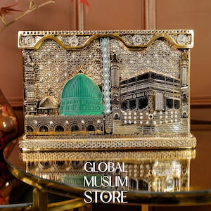Peut inclure: Une plaque décorative rectangulaire dorée présentant un relief détaillé de la Kaaba et du Dôme Vert. La surface est ornée de nombreux petits cristaux transparents et de motifs métalliques complexes, créant une finition texturée et réfléchissante.