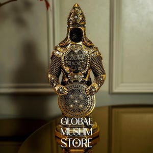 May include: A gold and black decorative figurine of a warrior holding a shield. The warrior is wearing a helmet and armor with intricate details. The shield is decorated with a geometric pattern. The base of the figurine reads "GLOBAL MUSLIM STORE".