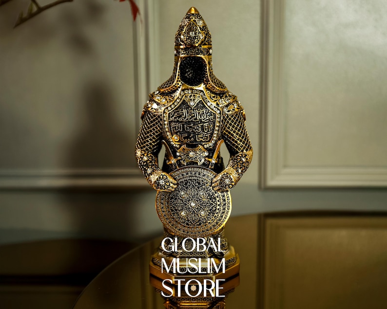 May include: A gold and black decorative figurine of a warrior holding a shield. The warrior is wearing a helmet and armor with intricate details. The shield is decorated with a geometric pattern. The base of the figurine reads "GLOBAL MUSLIM STORE".