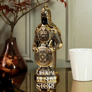 May include: A gold and black figurine of a warrior holding a shield with intricate designs. The warrior is wearing a helmet and armor with Arabic calligraphy. The base of the figurine reads "Global Muslim Store".