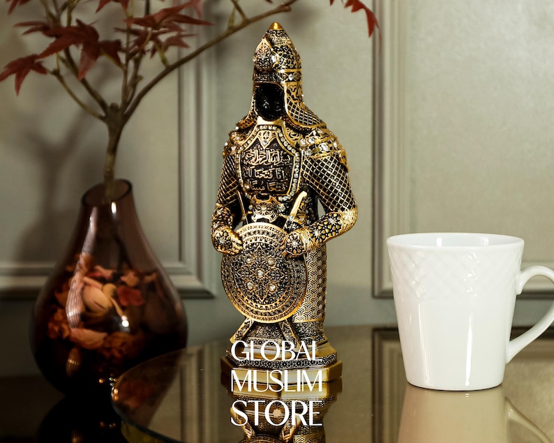 May include: A gold and black figurine of a warrior holding a shield with intricate designs. The warrior is wearing a helmet and armor with Arabic calligraphy. The base of the figurine reads "Global Muslim Store".