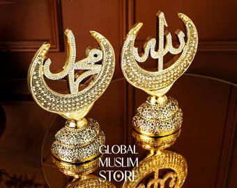 Pode incluir: Dois ornamentos de mesa decorativos de cor dourada em forma de meia-lua incrustados com cristais transparentes. Cada peça exibe caligrafia árabe e assenta numa base detalhada com padrão de filigrana. O texto GLOBAL MUSLIM STORE é visível.
