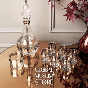 Peut inclure: Un décanteur en verre transparent avec un bouchon décoratif et des verres à liqueur assortis. Le décanteur et les verres présentent un motif de filigrane noir et or. L'image comprend également un vase avec des feuilles rouges et le texte "GLOBAL MUSLIM STORE".