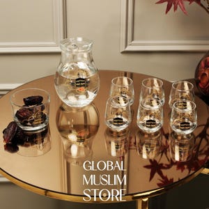 Op de afbeelding: Een heldere glazen karaf en zes bijpassende glazen met een gouden embleem, gerangschikt op een ronde, reflecterende tafel. Er staat ook een klein glas met dadels. De tafel heeft de tekst "GLOBAL MUSLIM STORE".