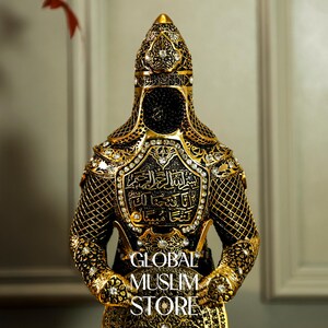 May include: A gold and black figurine of a warrior wearing a helmet and armor with intricate details. The armor is decorated with Arabic calligraphy and rhinestones. The figurine is standing with its hands clasped in front of it.