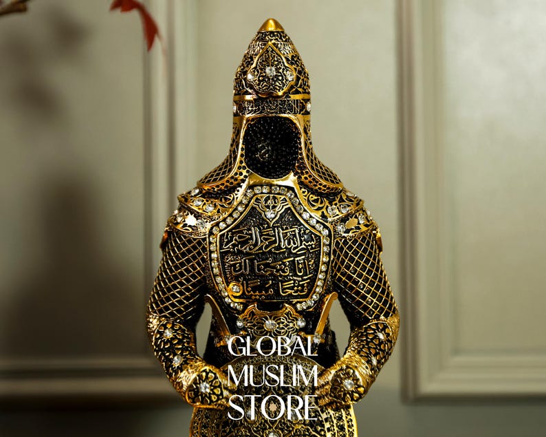 May include: A gold and black figurine of a warrior wearing a helmet and armor with intricate details. The armor is decorated with Arabic calligraphy and rhinestones. The figurine is standing with its hands clasped in front of it.