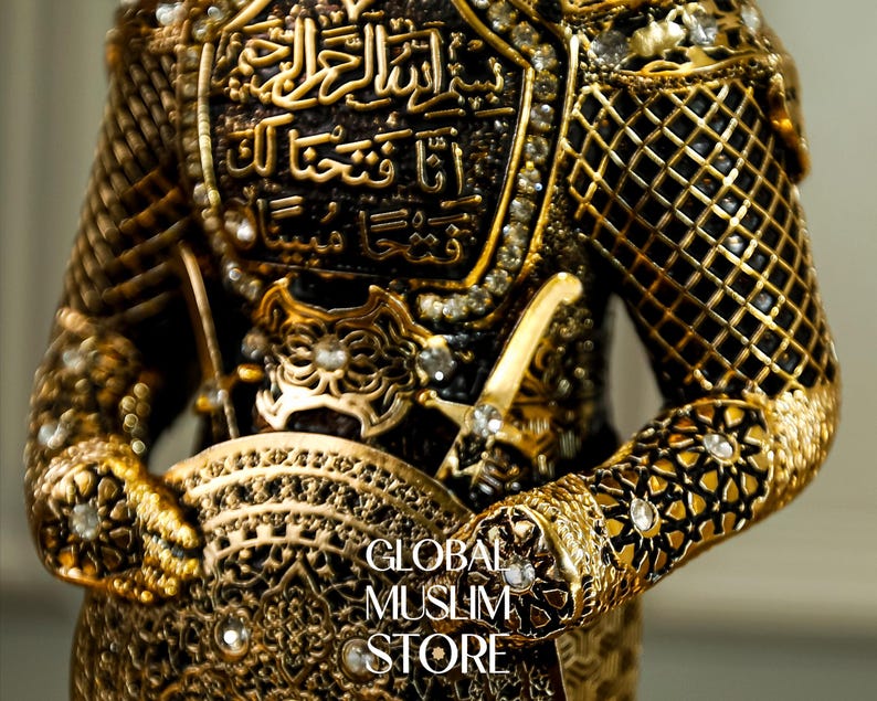 May include: A gold-colored figurine of a warrior wearing ornate armor with intricate details and holding a shield. The armor is decorated with rhinestones and Arabic calligraphy.