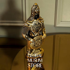 May include: A gold-colored figurine of a warrior wearing armor and holding a shield. The figurine is decorated with intricate details and has Arabic script on the chest plate. The figurine is on a base with the words "GLOBAL MUSLIM STORE" printed on it.