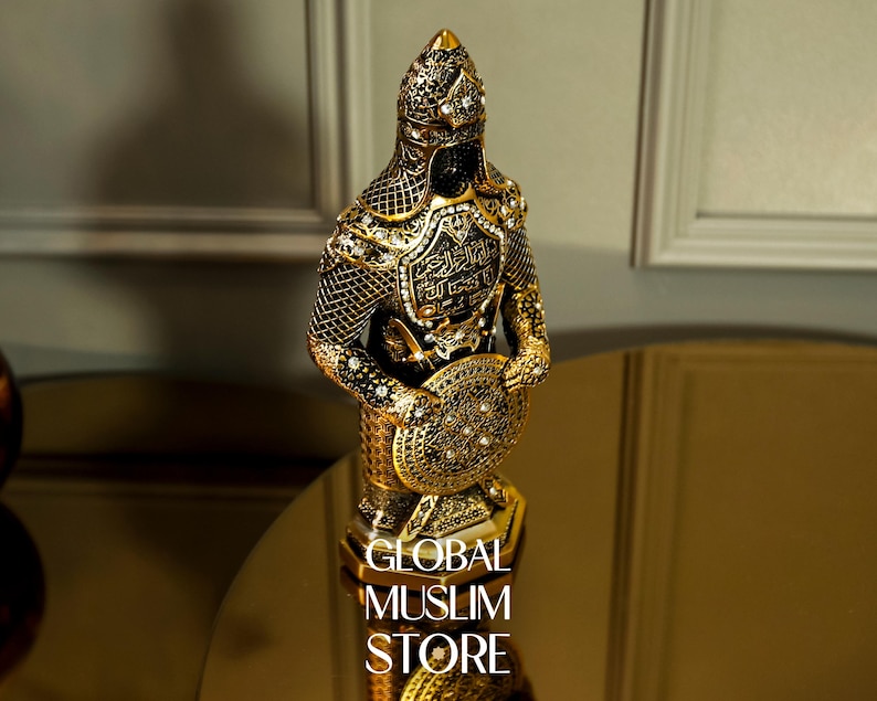 May include: A gold-colored figurine of a warrior wearing armor and holding a shield. The figurine is decorated with intricate details and has Arabic script on the chest plate. The figurine is on a base with the words "GLOBAL MUSLIM STORE" printed on it.