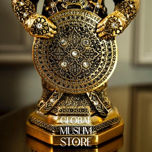 May include: A gold-colored metal figurine of a warrior holding a shield with intricate designs and rhinestones. The base of the figurine has the text "GLOBAL MUSLIM STORE".