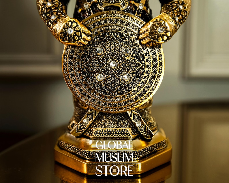 May include: A gold-colored metal figurine of a warrior holding a shield with intricate designs and rhinestones. The base of the figurine has the text "GLOBAL MUSLIM STORE".