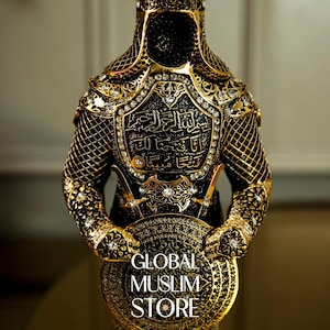 May include: A gold-colored metal figurine of a warrior wearing armor with intricate details and rhinestones. The warrior is holding a shield with the text "GLOBAL MUSLIM STORE" on it.