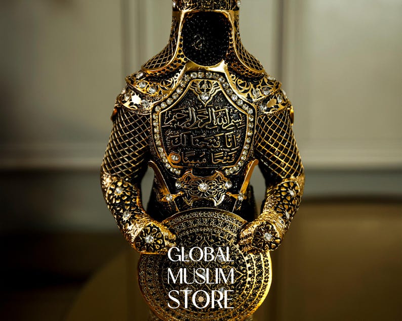 May include: A gold-colored metal figurine of a warrior wearing armor with intricate details and rhinestones. The warrior is holding a shield with the text "GLOBAL MUSLIM STORE" on it.