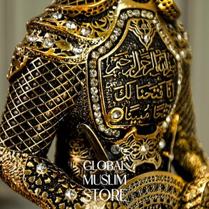 May include: A gold-colored figurine of a warrior wearing ornate armor with intricate details and rhinestones. The armor features Arabic calligraphy.