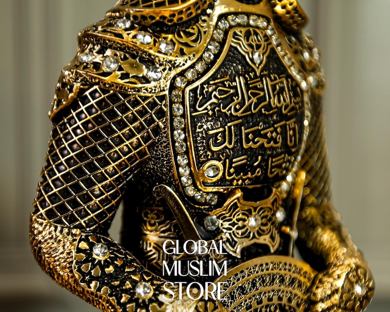 May include: A gold-colored figurine of a warrior wearing ornate armor with intricate details and rhinestones. The armor features Arabic calligraphy.