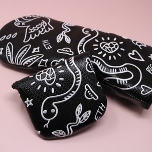 Puede incluir: Tres fundas para la cabeza de palo de golf negras con diseños bordados en blanco. Los diseños incluyen una serpiente, un corazón, un pájaro y las palabras "ETH" y "BTC".