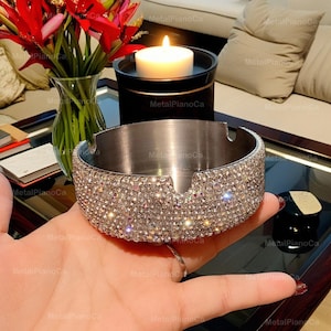 Könnte beinhalten: Ein silberner Aschenbecher mit einer runden, flachen Schale und einem breiten Rand, der mit funkelnden, klaren Strasssteinen besetzt ist.