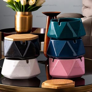May include: Four ceramic ashtrays with geometric designs. The ashtrays are stacked in a pyramid shape. The top ashtray is teal green, the middle ashtray is dark blue, and the bottom ashtray is pink. The ashtray on the left is black with a white base and a wooden lid.