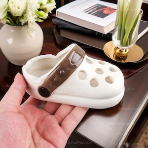 Könnte beinhalten: Weißer Aschenbecher aus Keramik in Form eines Clogs mit braunem Riemendetail. Der Aschenbecher hat eine runde Öffnung und mehrere Löcher oben.