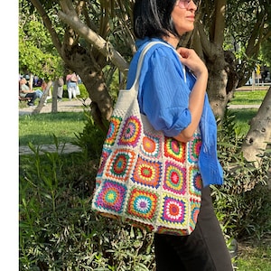 May include: A white crochet tote bag with colorful square patches. The bag has a large, open top and is made with a thick, textured yarn. The patches are made with a variety of colors, including red, orange, yellow, green, blue, and purple.