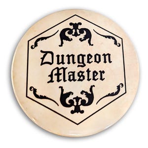 Op de afbeelding: Een ronde, crèmekleurige knop met een zwart zeshoekig ontwerp. Het ontwerp bevat een gestileerde inscriptie met de tekst "Dungeon Master" en decoratieve versieringen.