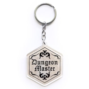 Puede incluir: Un llavero hexagonal transparente con las palabras "Dungeon Master" en una fuente decorativa. El llavero tiene un anillo y una cadena de color plateado. El diseño es en blanco y negro, con detalles de borde ornamentados.
