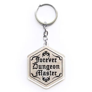 Può includere: Un portachiavi esagonale trasparente con la scritta "Forever Dungeon Master" in nero, con lettere ornate. Il portachiavi ha un portachiavi e una catena argentati, perfetto per i fan dei giochi di ruolo da tavolo.