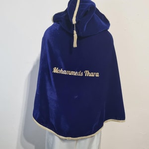 May include: A blue velvet hooded cape with gold trim and a gold tassel. The cape has the name "Mohammeds Thara" embroidered on the front in gold thread.