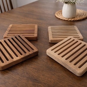 May include: Four square wooden trivets with a slatted design are arranged on a dark wood table. Two trivets are a lighter wood color, and two are a darker wood color. A vase of flowers sits in the background.