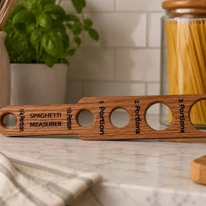 May include: Wooden spaghetti measurer with holes for 1/4, 1/2, 1, 2, and 3 portions. The text "SPAGHETTI MEASURER" is engraved on the wood. The kitchen tool is made of light brown wood and sits on a marble countertop.