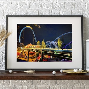 May include: A framed photograph of a roller coaster at an amusement park at night. The roller coaster is lit up with blue lights and the track is visible against the dark sky.