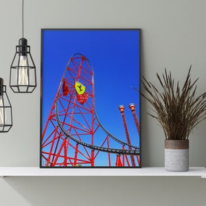 May include: A red and black roller coaster with a yellow Ferrari logo against a bright blue sky. The roller coaster is in a park setting with green trees and a white shelf in the foreground.