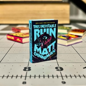 Op de afbeelding: Een miniatuurboek getiteld "This Inevitable Ruin" van Matt Dinniman. Het boek heeft een zwarte kaft met turquoise en roze tekst en een turquoise rug. Andere miniatuurboeken staan op de achtergrond.