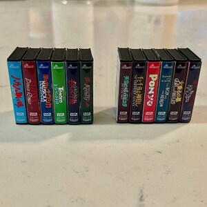 May include: Two sets of miniature VHS tapes, each with several colourful movie titles. The tapes are arranged in two neat rows, with titles like "Spirited Away" and "My Neighbour Totoro" visible. The tapes have a vintage aesthetic.