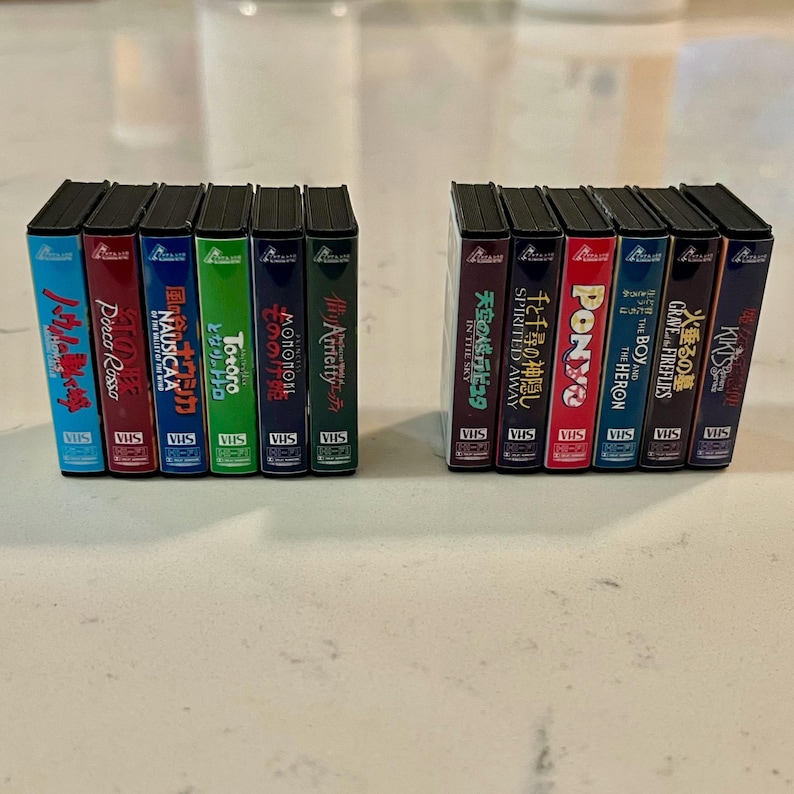 May include: Two sets of miniature VHS tapes, each with several colourful movie titles. The tapes are arranged in two neat rows, with titles like "Spirited Away" and "My Neighbour Totoro" visible. The tapes have a vintage aesthetic.