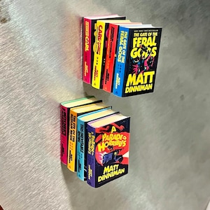 Op de afbeelding: Miniatuur boeken set met titels als "The Gate of the Feral Gods" en "A Parade of Horribles" van Matt Dinniman. De boeken hebben kleurrijke ruggen en gedetailleerde cover illustraties, ideaal voor verzamelaars.