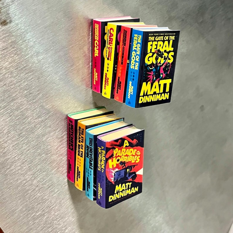 Op de afbeelding: Miniatuur boeken set met titels als "The Gate of the Feral Gods" en "A Parade of Horribles" van Matt Dinniman. De boeken hebben kleurrijke ruggen en gedetailleerde cover illustraties, ideaal voor verzamelaars.