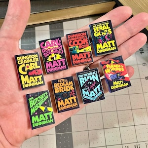 Op de afbeelding: Een verzameling miniatuurboekreplica's met kleurrijke covers. Titels zijn onder meer "Dungeon Crawler", "Carl's Doomsday Scenario" en "The Feral Gods". Elk boek toont de naam van de auteur, Matt Dinniman, in een vetgedrukte letter.